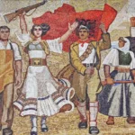 Une fresque de style socialiste représentant un groupe d’agitateurs albanais en marche, avec une figure féminine centrale levant un fusil. Les personnages portent des vêtements traditionnels et de travail, symbolisant les différentes classes sociales unies, avec en arrière-plan un drapeau rouge arborant l’aigle bicéphale, symbole de l’Albanie. Les agitateurs avancent avec des expressions déterminées, mettant en valeur l’esprit révolutionnaire et la lutte pour la liberté.