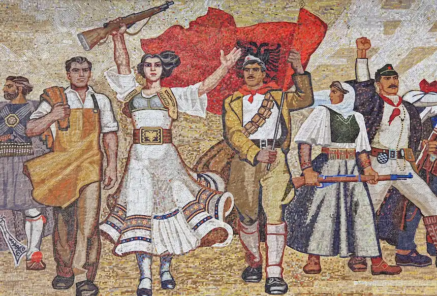 Une fresque de style socialiste représentant un groupe d’agitateurs albanais en marche, avec une figure féminine centrale levant un fusil. Les personnages portent des vêtements traditionnels et de travail, symbolisant les différentes classes sociales unies, avec en arrière-plan un drapeau rouge arborant l’aigle bicéphale, symbole de l’Albanie. Les agitateurs avancent avec des expressions déterminées, mettant en valeur l’esprit révolutionnaire et la lutte pour la liberté.