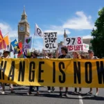 a climate activism protest, featuring demonstrators holding banners and signs with messages like "Change is Now" and "Just Stop Oil." The setting includes prominent landmarks, suggesting the protest's visibility in a public or political hub.