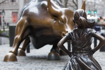A bronze statue of the "Fearless Girl" stands confidently with hands on her hips, facing the iconic "Charging Bull" of Wall Street. The image symbolizes the tension between corporate power and evolving values of Corporate Social Responsibility, inclusivity, and sustainability in modern business practices.