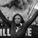 Una mujer levantando una bengala de humo durante una protesta, rodeada de manifestantes con pancartas, simbolizando la lucha continua contra la opresión y la desigualdad. Esta poderosa imagen refleja los principios de la sociología feminista.