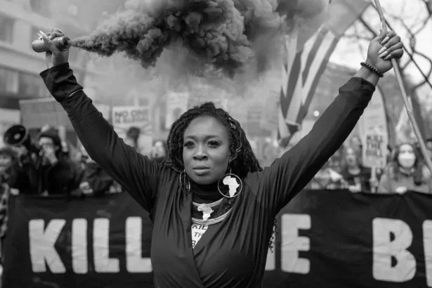 A woman raising a smoke flare during a protest, surrounded by demonstrators holding signs, symbolizing the ongoing fight against oppression and inequality. This powerful image reflects the principles of feminist sociology.