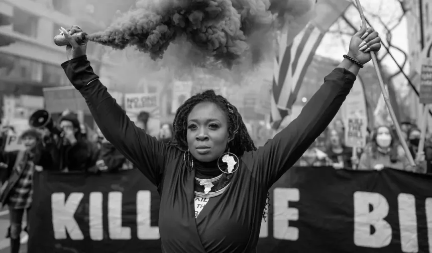 A woman raising a smoke flare during a protest, surrounded by demonstrators holding signs, symbolizing the ongoing fight against oppression and inequality. This powerful image reflects the principles of feminist sociology.
