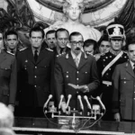 A black and white photograph of military perpetrators during Argentina's 1976 coup d'état. Prominent military leaders, including Jorge Rafael Videla (center), stand at attention in uniforms behind multiple microphones. Videla reads from a paper, symbolizing the Junta Militar's formal announcement of their seizure of power. Other perpetrators, Emilio Massera (left) and Orlando Ramón Agosti (right), flank Videla, representing the Argentine Army, Navy, and Air Force, key orchestrators of state terrorism during the dictatorship.