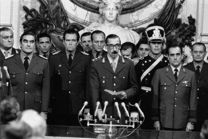 A black and white photograph of military perpetrators during Argentina's 1976 coup d'état. Prominent military leaders, including Jorge Rafael Videla (center), stand at attention in uniforms behind multiple microphones. Videla reads from a paper, symbolizing the Junta Militar's formal announcement of their seizure of power. Other perpetrators, Emilio Massera (left) and Orlando Ramón Agosti (right), flank Videla, representing the Argentine Army, Navy, and Air Force, key orchestrators of state terrorism during the dictatorship.