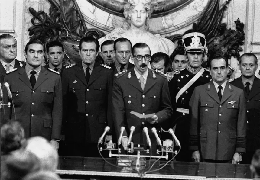 A black and white photograph of military perpetrators during Argentina's 1976 coup d'état. Prominent military leaders, including Jorge Rafael Videla (center), stand at attention in uniforms behind multiple microphones. Videla reads from a paper, symbolizing the Junta Militar's formal announcement of their seizure of power. Other perpetrators, Emilio Massera (left) and Orlando Ramón Agosti (right), flank Videla, representing the Argentine Army, Navy, and Air Force, key orchestrators of state terrorism during the dictatorship.