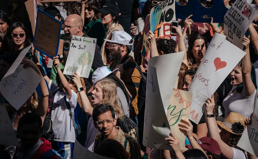 Una multitud de manifestantes sosteniendo varios carteles hechos a mano durante una protesta climática. Uno de los carteles pregunta: "¿Qué verde ves?", mostrando un árbol junto a dinero, mientras otros expresan preocupaciones sobre la justicia ambiental. Esta imagen captura la urgencia de abordar la violencia ambiental como un tema global de derechos humanos.