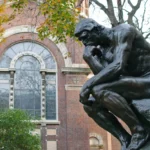 La escultura El Pensador de Rodin en la Universidad de Columbia, rodeada de follaje otoñal y situada frente a un edificio histórico de ladrillo con grandes ventanas arqueadas. La obra simboliza la soledad y la reflexión profunda.