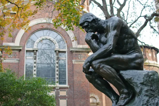 La sculpture Le Penseur de Rodin à l'Université Columbia, entourée de feuillage automnal et située devant un bâtiment historique en briques avec de grandes fenêtres en arc. L'œuvre symbolise la solitude et la réflexion profonde.