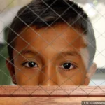 Un enfant regarde à travers une barrière en treillis métallique, avec une expression sombre, symbolisant l'impact des politiques de séparation familiale à la frontière. Les figures en arrière-plan sont floues. Photo créditée à l'Office des douanes et de la protection des frontières des États-Unis.