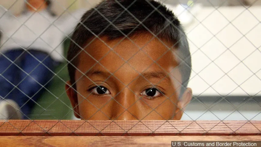 A young boy looks through a mesh wire barrier, with a somber expression, symbolizing the impact of family separation policies at the border. Background figures are blurred. Photo credited to U.S. Customs and Border Protection.