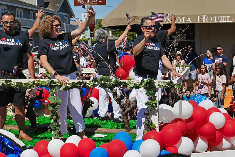 Participants in a parade wearing shirts that read 'We Are All Immigrants,' holding small American flags amidst colorful decorations, symbolizing support for inclusive immigration policies.