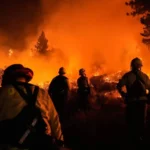 Injusticia ambiental: Un grupo de bomberos enfrenta un incendio forestal descomunal, con sus siluetas iluminadas por el intenso resplandor naranja de las llamas.