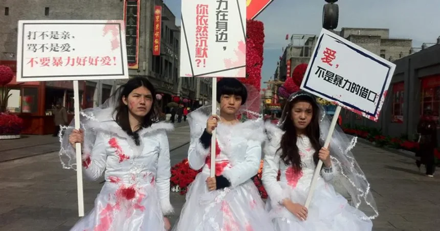 Mascarada digital. Tres activistas feministas chinas vestidas de novias, con manchas rojas que asemejan sangre, sostienen carteles de protesta contra la violencia de género en una calle concurrida. Sus expresiones son serias y desafiantes, simbolizando la resistencia contra la censura y la represión estatal. La escena representa una forma creativa de activismo feminista y queer en China.