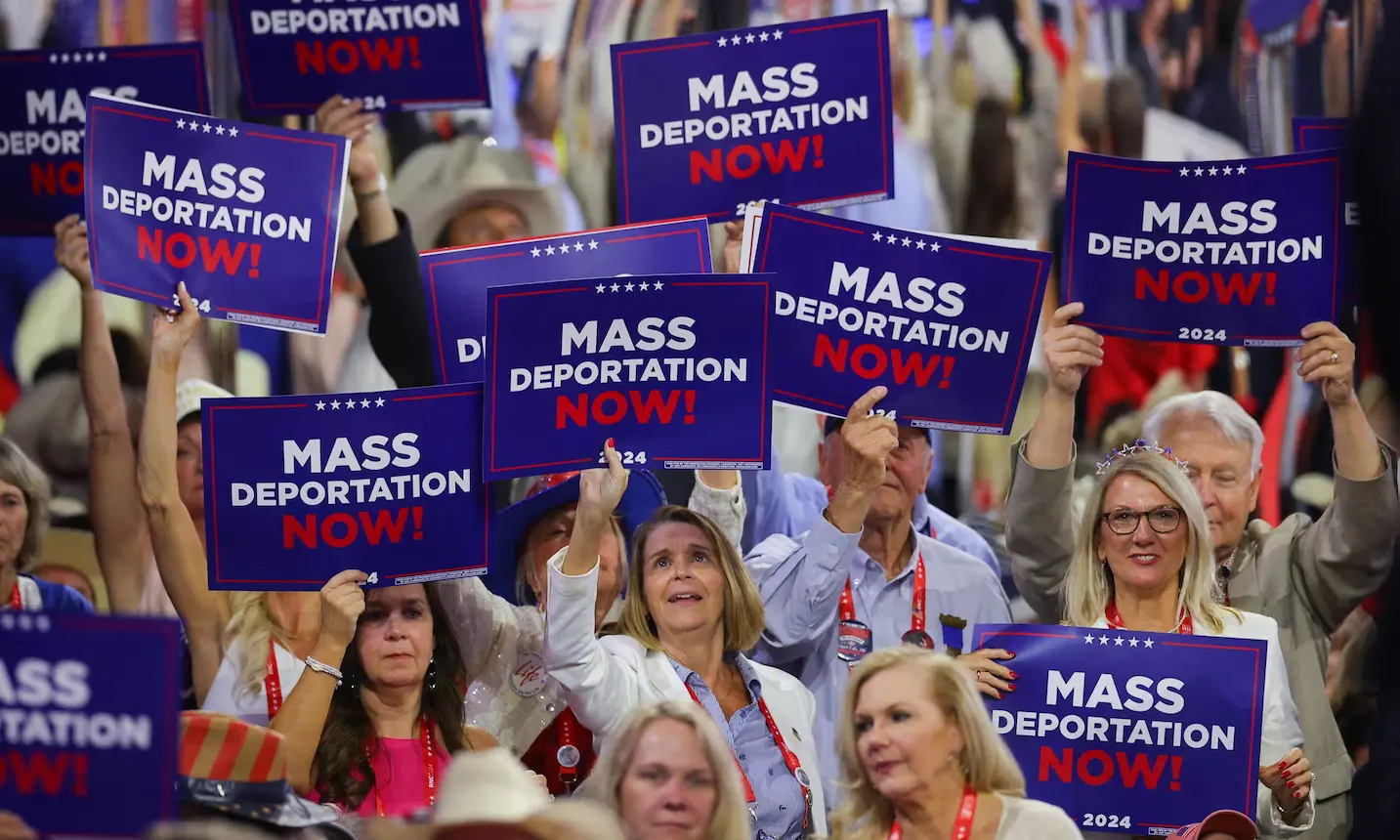 Nativism in action: A crowd of people holding up blue and red signs that read 'MASS DEPORTATION NOW!' in bold white and red letters. The signs have stars and the year '2024' printed on them, indicating a political event or rally. The individuals appear engaged, some smiling, while others have serious expressions, reflecting strong sentiments around the topic of immigration.