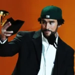 Bad Bunny, wearing a green baseball cap and a black suit, holds up a Grammy award while speaking at the microphone. His working-class aesthetic, often reflected in his music and performances, raises questions about labor, authenticity, and pop stardom.