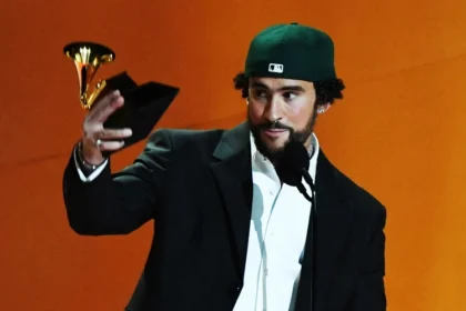 Bad Bunny, con una gorra de béisbol verde y un traje negro, sostiene un premio Grammy mientras habla ante el micrófono. Su estética de clase trabajadora, reflejada con frecuencia en su música y presentaciones, plantea interrogantes sobre el trabajo, la autenticidad y la fama en el pop.