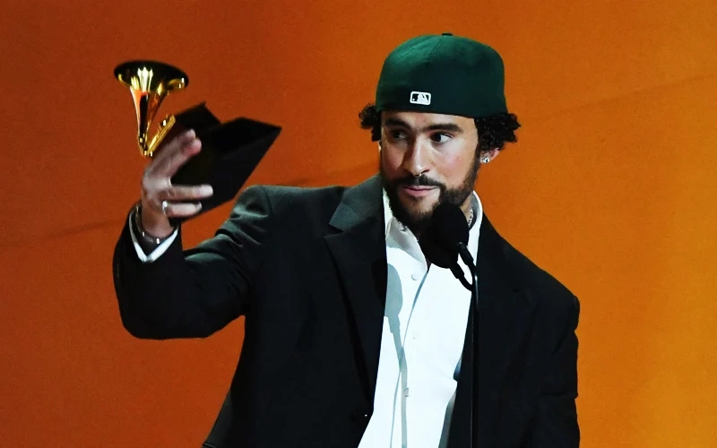 Bad Bunny, wearing a green baseball cap and a black suit, holds up a Grammy award while speaking at the microphone. His working-class aesthetic, often reflected in his music and performances, raises questions about labor, authenticity, and pop stardom.