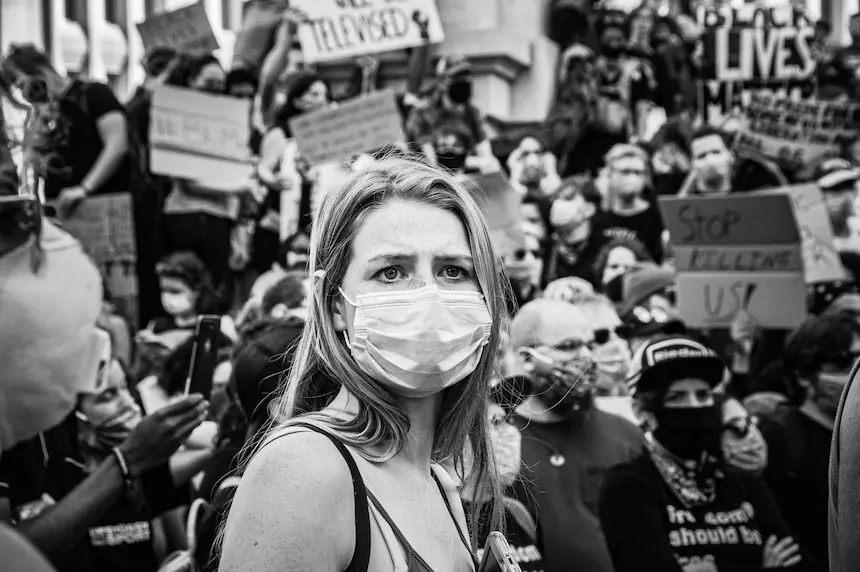 Black and white photograph of a masked woman in the foreground of a protest, surrounded by demonstrators holding signs advocating for racial justice and social change. The image captures the tension, urgency, and emotional weight of civil unrest and political activism in a divided society.