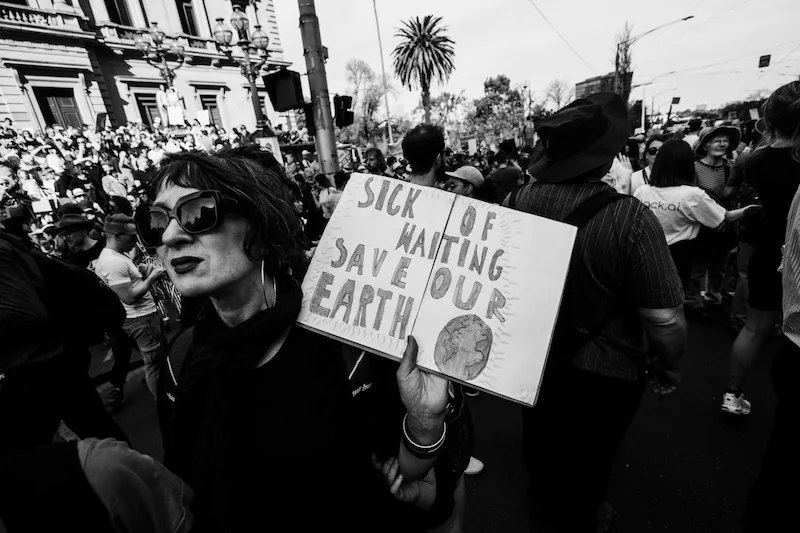 Disintegrating Societies: A black and white photograph of a climate protest featuring a woman holding a sign that reads 'Sick of Waiting, Save Our Earth.' The image captures the urgency of environmental activism amid growing societal and political fragmentation.