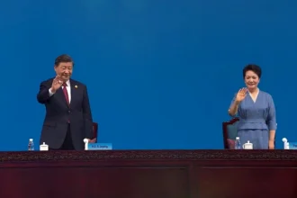 Love: Xi Jinping and Peng Liyuan wave from behind a long wooden table at an official event, standing against a plain blue background.
