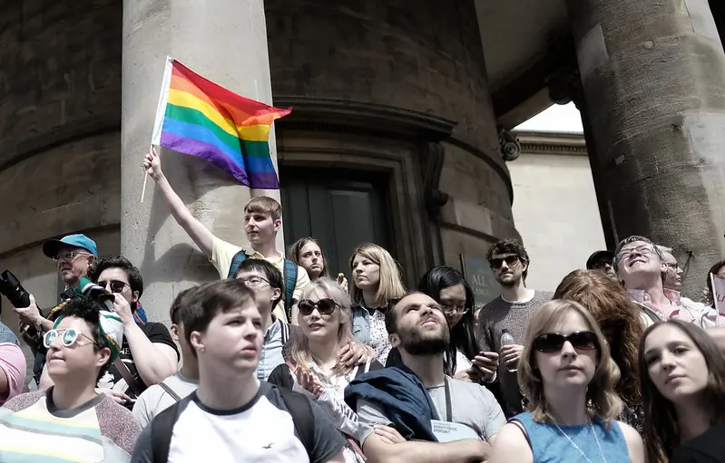 Un participant lève le drapeau de la Fierté lors d'un rassemblement public, affirmant la présence et les droits des personnes LGBTQ+ face à la marginalisation continue. La visibilité dans l'espace public reste une forme cruciale de voix politique et de résistance. Photo par Alisdare Hickson (CC BY-SA).