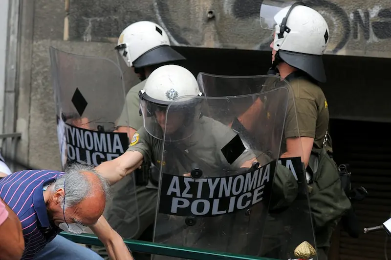 Riot police confront an elderly protester during anti-austerity demonstrations in Athens—an emblematic scene of neoliberalism’s social fallout in Southern Europe.