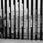 Une silhouette solitaire fait face à la clôture frontalière à la limite entre les États-Unis et le Mexique. L’image évoque des questions d’exclusion, de visibilité et du coût humain des frontières nationales — des thèmes au cœur des débats contemporains sur la politique migratoire. Photo de Max Bohme.