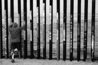 Una figura solitaria se enfrenta a la valla fronteriza en la división entre Estados Unidos y México. La imagen evoca cuestiones de exclusión, visibilidad y el costo humano de las fronteras nacionales, todos ellos temas centrales en los debates contemporáneos sobre política migratoria. Foto de Max Bohme.