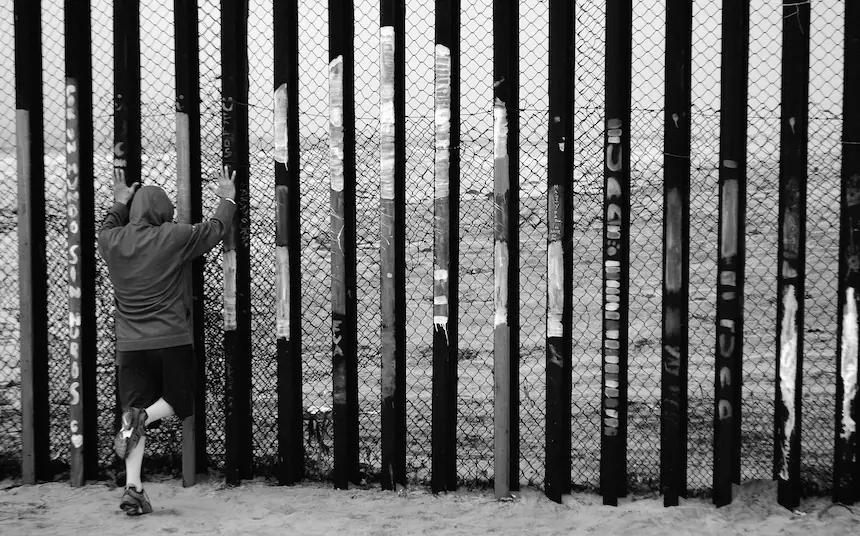 A solitary figure faces the border fence at the U.S.–Mexico divide. The image evokes questions of exclusion, visibility, and the human cost of national boundaries — all central themes in contemporary immigration policy debates.