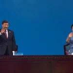 Love: Xi Jinping and Peng Liyuan wave from behind a long wooden table at an official event, standing against a plain blue background.