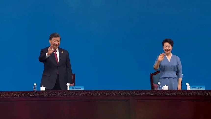 Love: Xi Jinping and Peng Liyuan wave from behind a long wooden table at an official event, standing against a plain blue background.