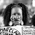 Una mujer negra sostiene una pancarta con los nombres de víctimas de la violencia policial durante una protesta en Estados Unidos. Su mascarilla lleva los rostros de personas negras asesinadas por la policía, entre ellas George Floyd. Foto de Lorie Shaull (CC BY).