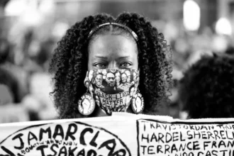 A Black woman holds a banner listing victims of police violence during a protest in the United States. Her mask features the faces of Black individuals killed by police, including George Floyd.