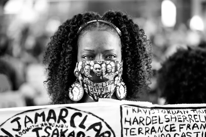 Une femme noire tient une bannière énumérant les victimes de violences policières lors d’une manifestation aux États-Unis. Son masque arbore les visages de personnes noires tuées par la police, dont George Floyd. Photo de Lorie Shaull (CC BY).