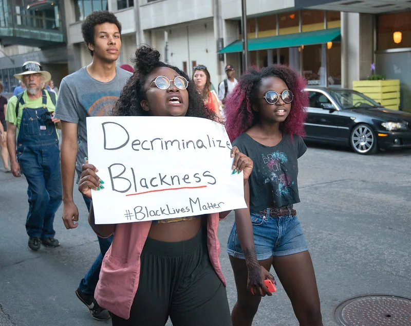 De jeunes femmes noires manifestent en portant une pancarte indiquant « Décriminaliser la négritude », reprenant une revendication centrale du mouvement Black Lives Matter contre la police racialisée. Photo de Fibonacci Blue (CC BY).