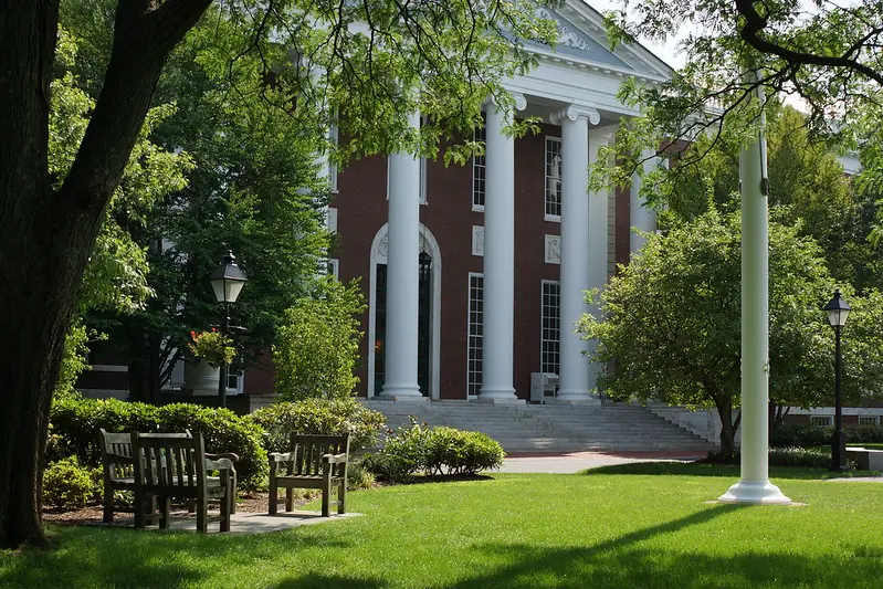 Harvard Business School building with white columns and red brick façade, surrounded by trees and benches on a quiet green lawn. (Photo credit: Michael A. Herzog, CC BY)