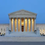 Supreme Court of the United States building at dusk in Washington, D.C., illuminated against a clear evening sky. Photo by Joe Ravi, CC BY-SA.