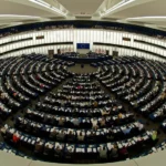 National Security State discussion: The circular plenary chamber of the European Parliament in session, filled with members, with EU flags and emblem at the front.