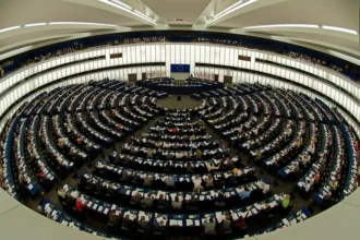 National Security State discussion: The circular plenary chamber of the European Parliament in session, filled with members, with EU flags and emblem at the front.