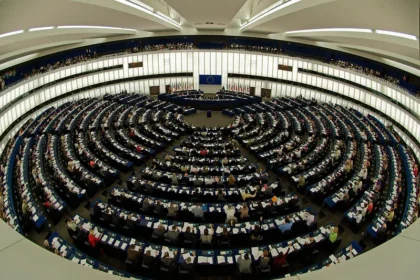 National Security State discussion: The circular plenary chamber of the European Parliament in session, filled with members, with EU flags and emblem at the front.