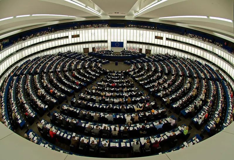 National Security State discussion: The circular plenary chamber of the European Parliament in session, filled with members, with EU flags and emblem at the front.