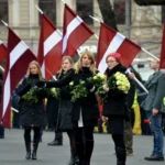 A public ceremony in Latvia where participants carry national flags and white flowers, illustrating the rituals through which nationhood is symbolically reproduced in public life.