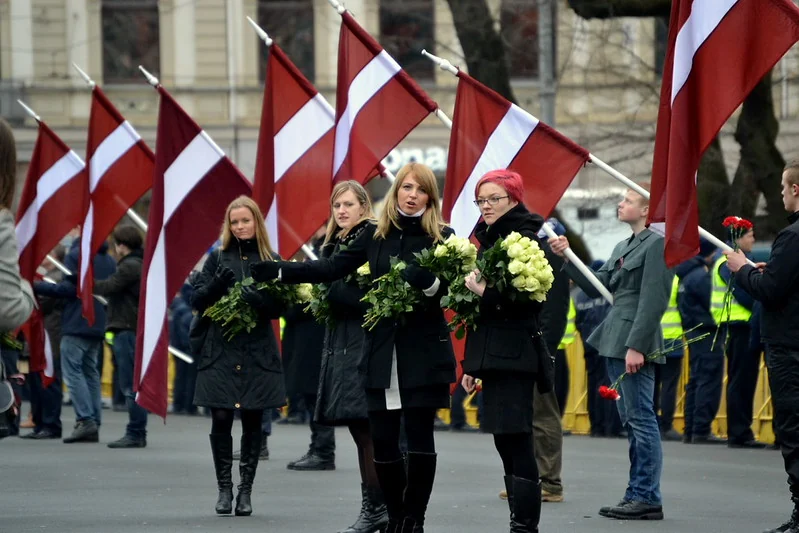A public ceremony in Latvia where participants carry national flags and white flowers, illustrating the rituals through which nationhood is symbolically reproduced in public life.