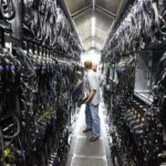 Two technicians confer in a narrow server aisle inside a data center, flanked by racks and dense cabling that illustrate the physical infrastructure now at the center of the AI backlash. Photo by Robert Scoble ( CC BY)