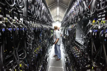 Two technicians confer in a narrow server aisle inside a data center, flanked by racks and dense cabling that illustrate the physical infrastructure now at the center of the AI backlash. Photo by Robert Scoble ( CC BY)