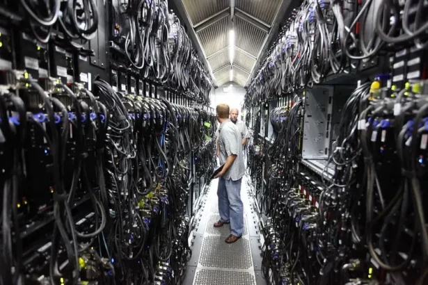 Deux techniciens s’entretiennent dans une allée de serveurs étroite au sein d’un centre de données, entourés de baies et d’un câblage dense qui illustrent l’infrastructure physique désormais au cœur de la contestation de l’IA. Photo de Robert Scoble (CC BY)