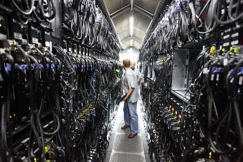 Two technicians confer in a narrow server aisle inside a data center, flanked by racks and dense cabling that illustrate the physical infrastructure now at the center of the AI backlash. Photo by Robert Scoble ( CC BY)