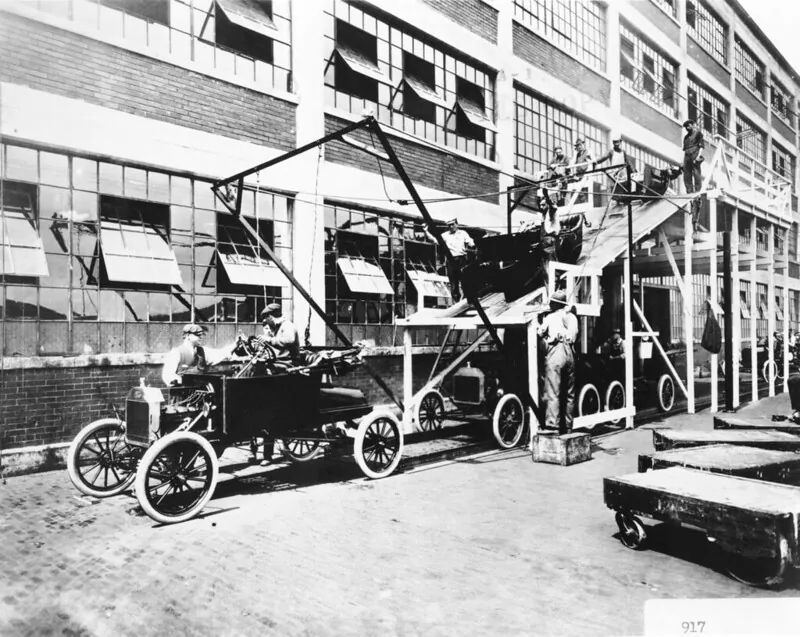 Workers assemble a Ford Model T outside a factory using an overhead Power rail system